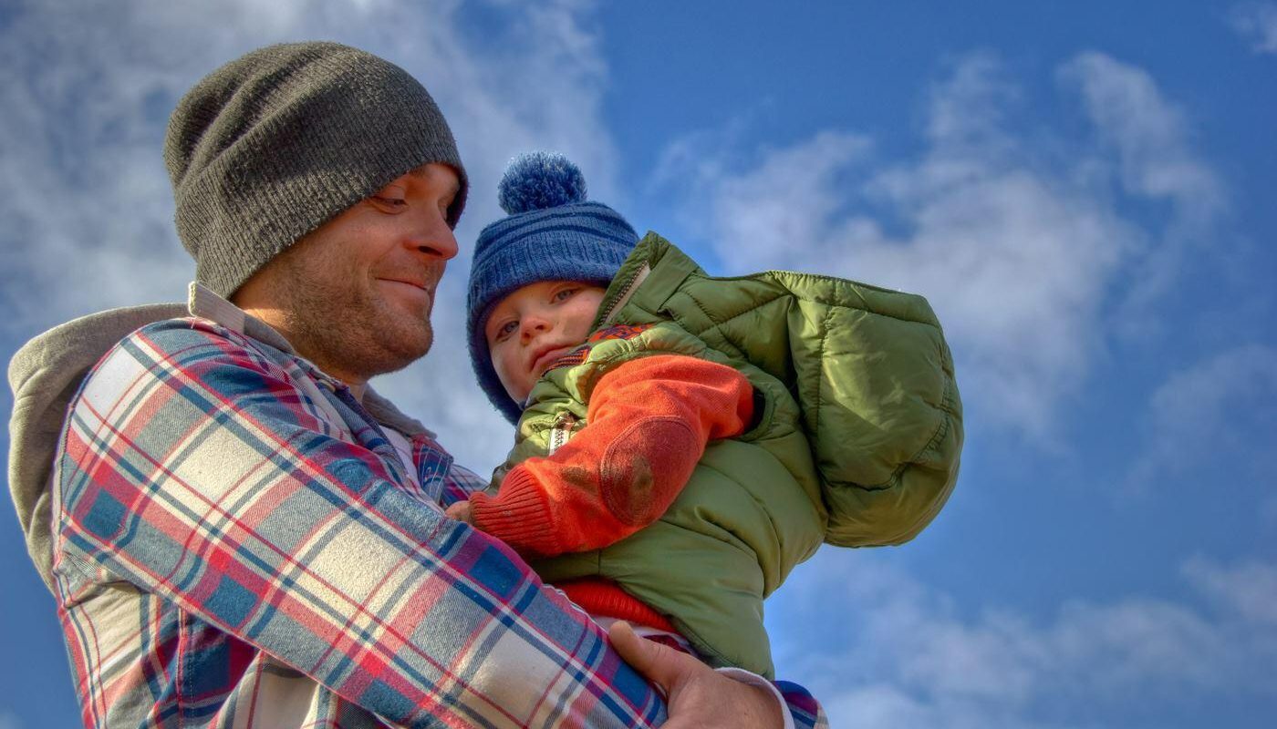 Man in a beanie holding his son dressed warmly for winter with a blue sky background